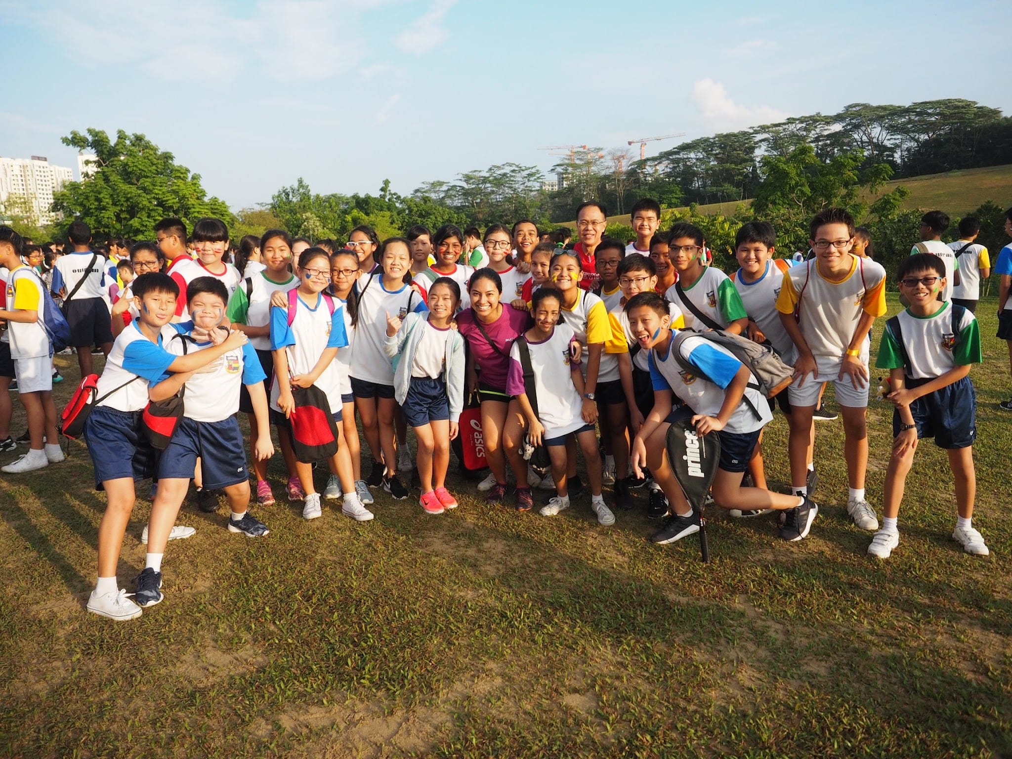 A large group of students in school sports uniforms pose together smiling on a grass field with trees in the background.
