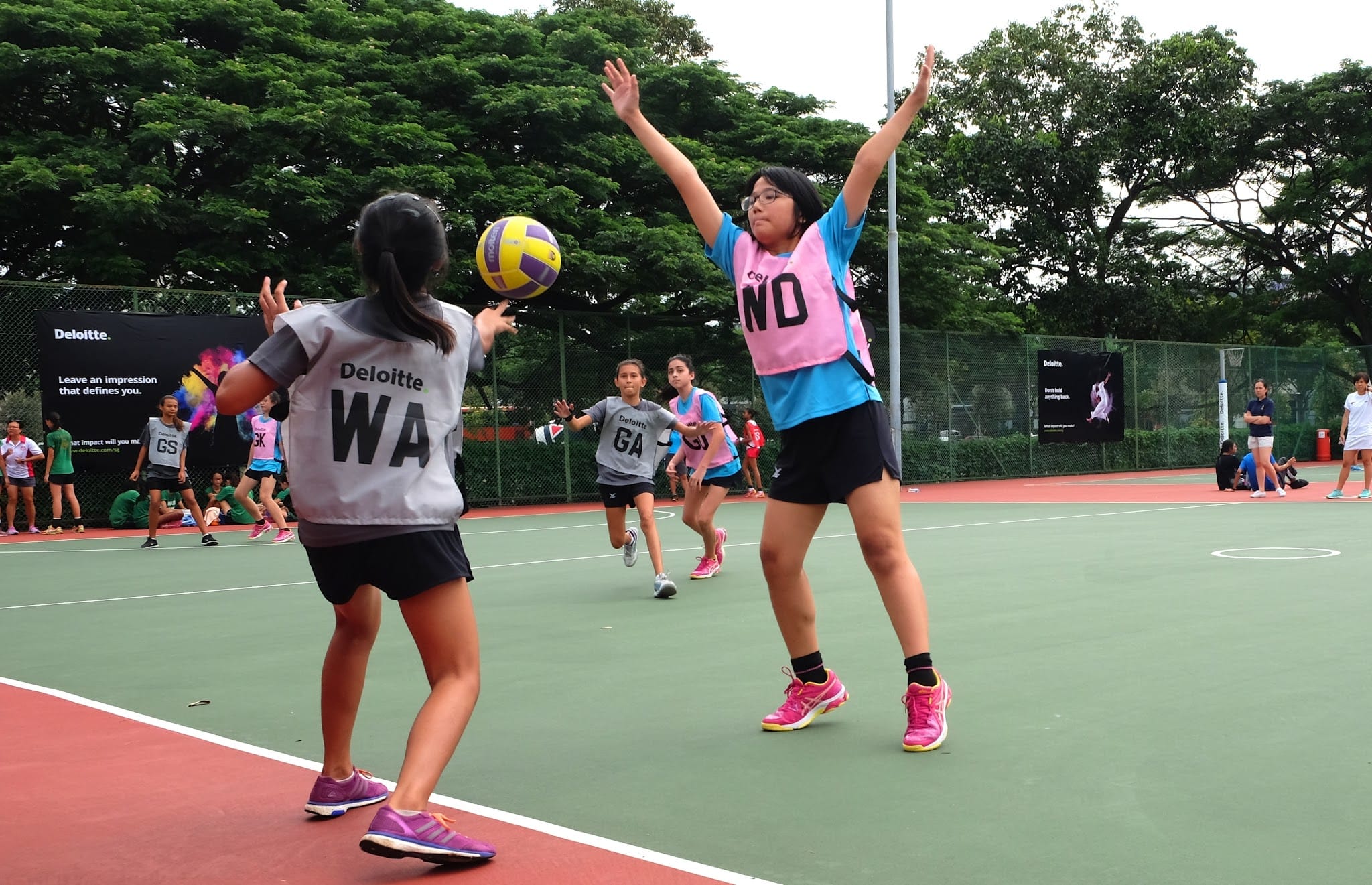 Girls playing netball outdoors, one jumping with arms raised to block a yellow and purple ball from a teammate.