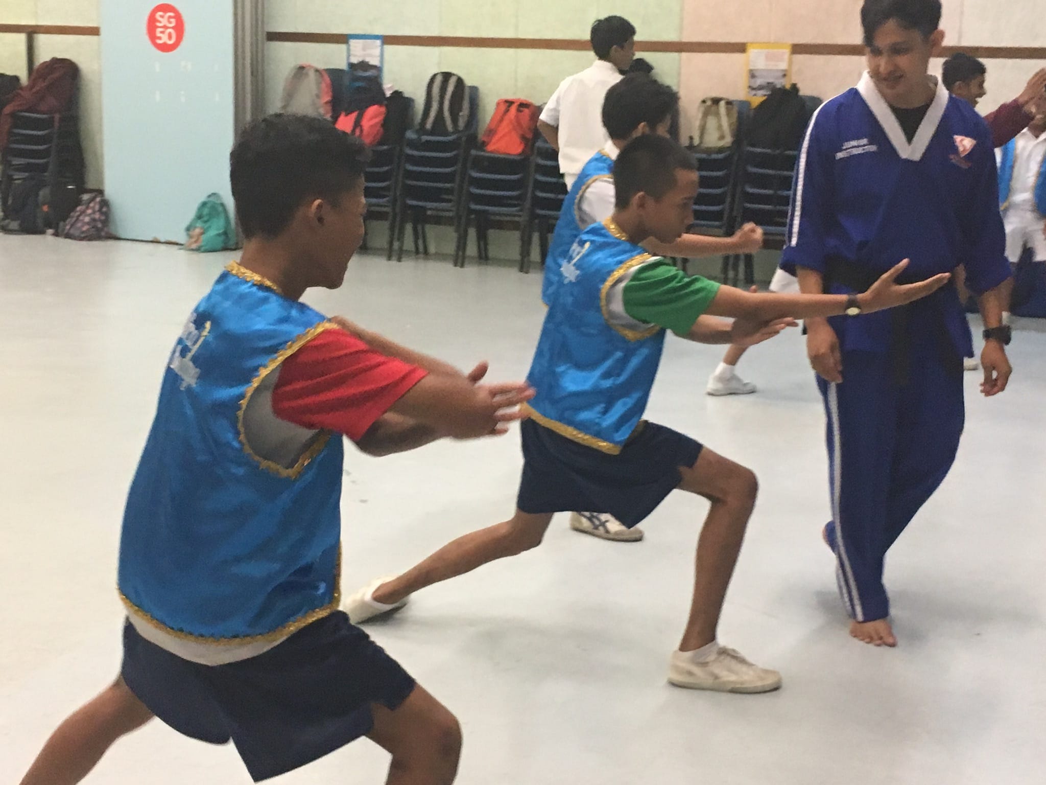 Two boys in blue vests practice martial arts moves in a hall while an instructor in a blue uniform watches.