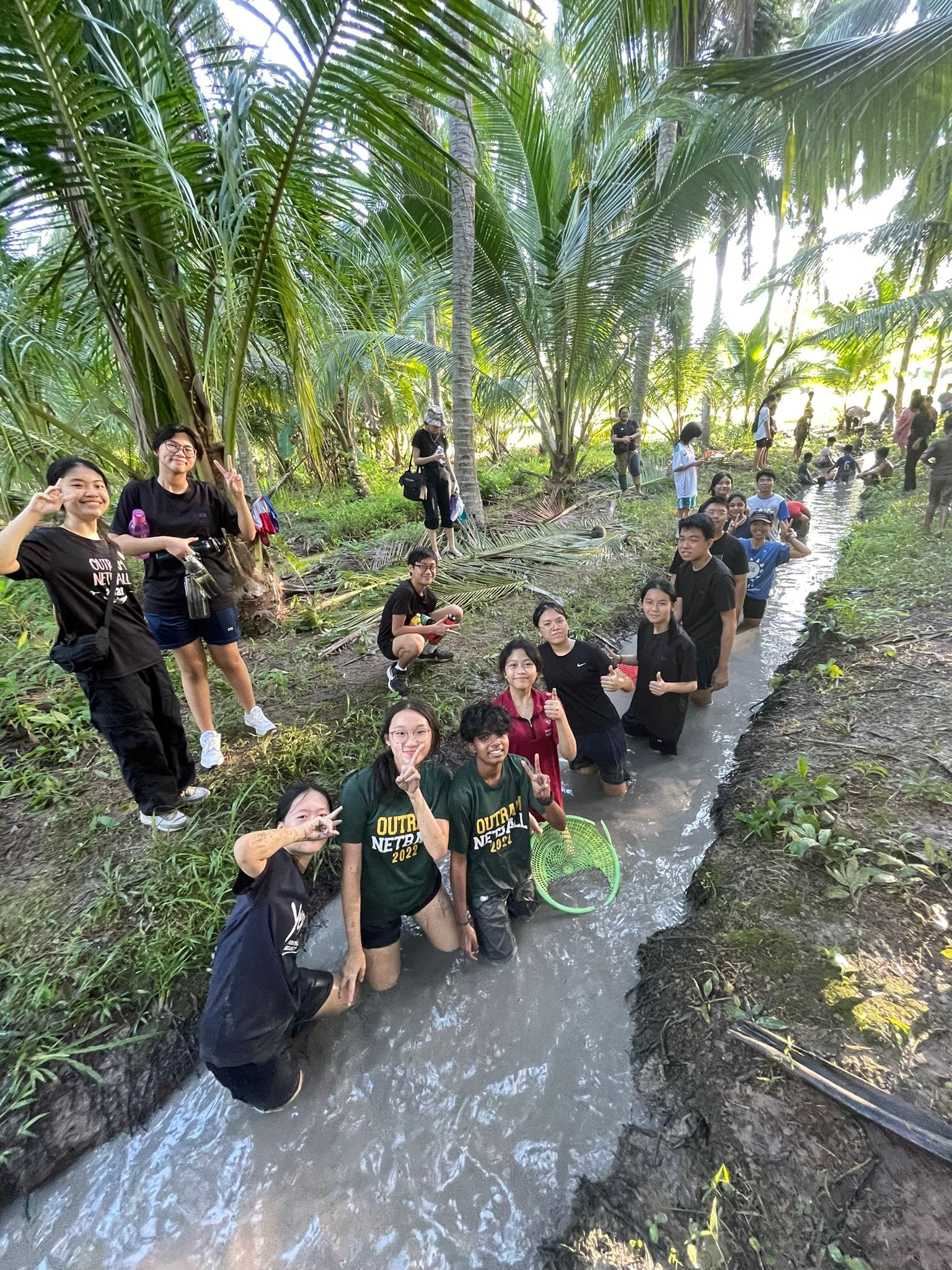 A group of smiling young people standing and kneeling in a muddy canal surrounded by palm trees, posing with peace signs.