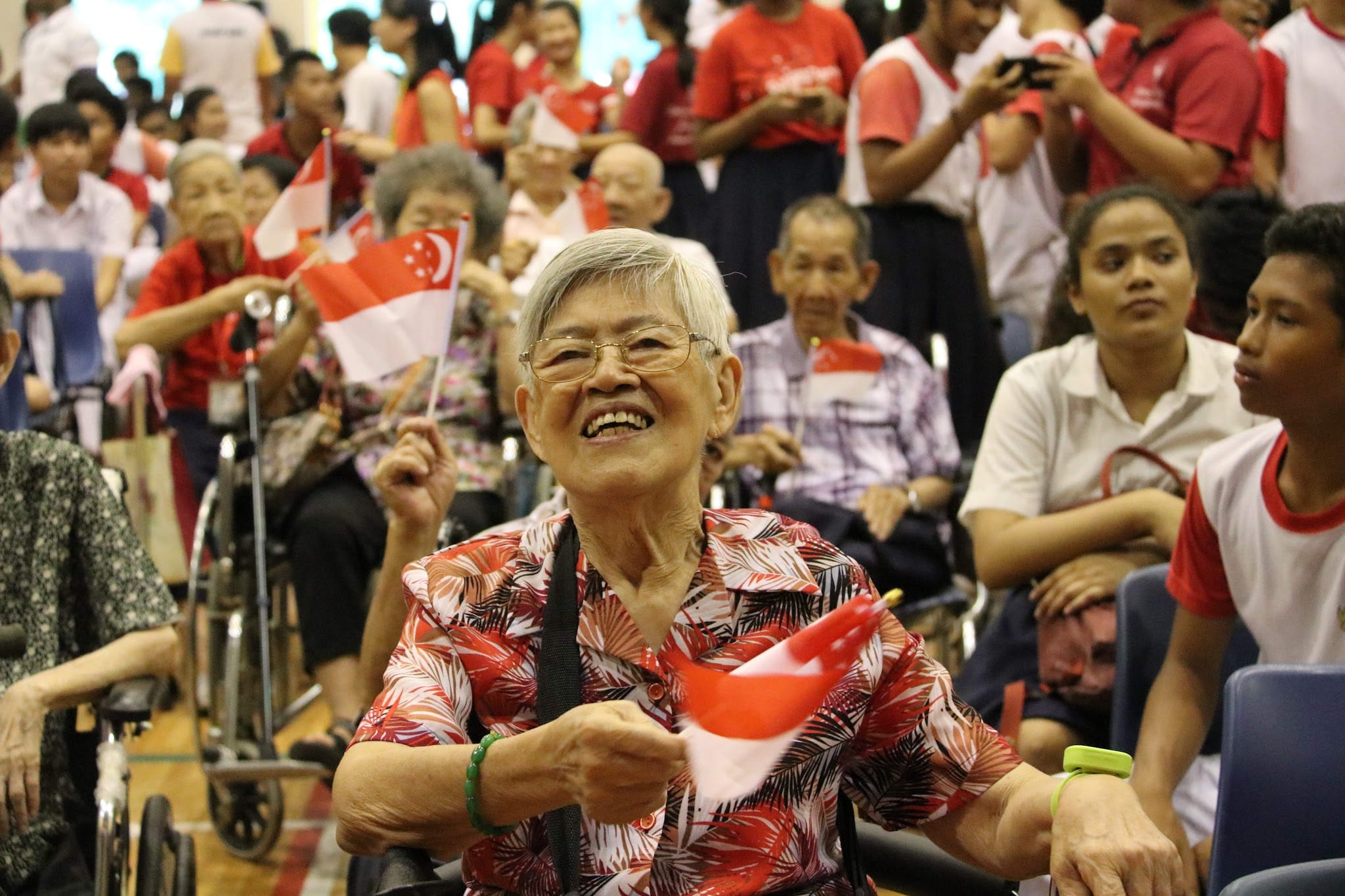 Elderly woman smiling and holding a Singapore flag at a crowded National Day event with people in red and white.