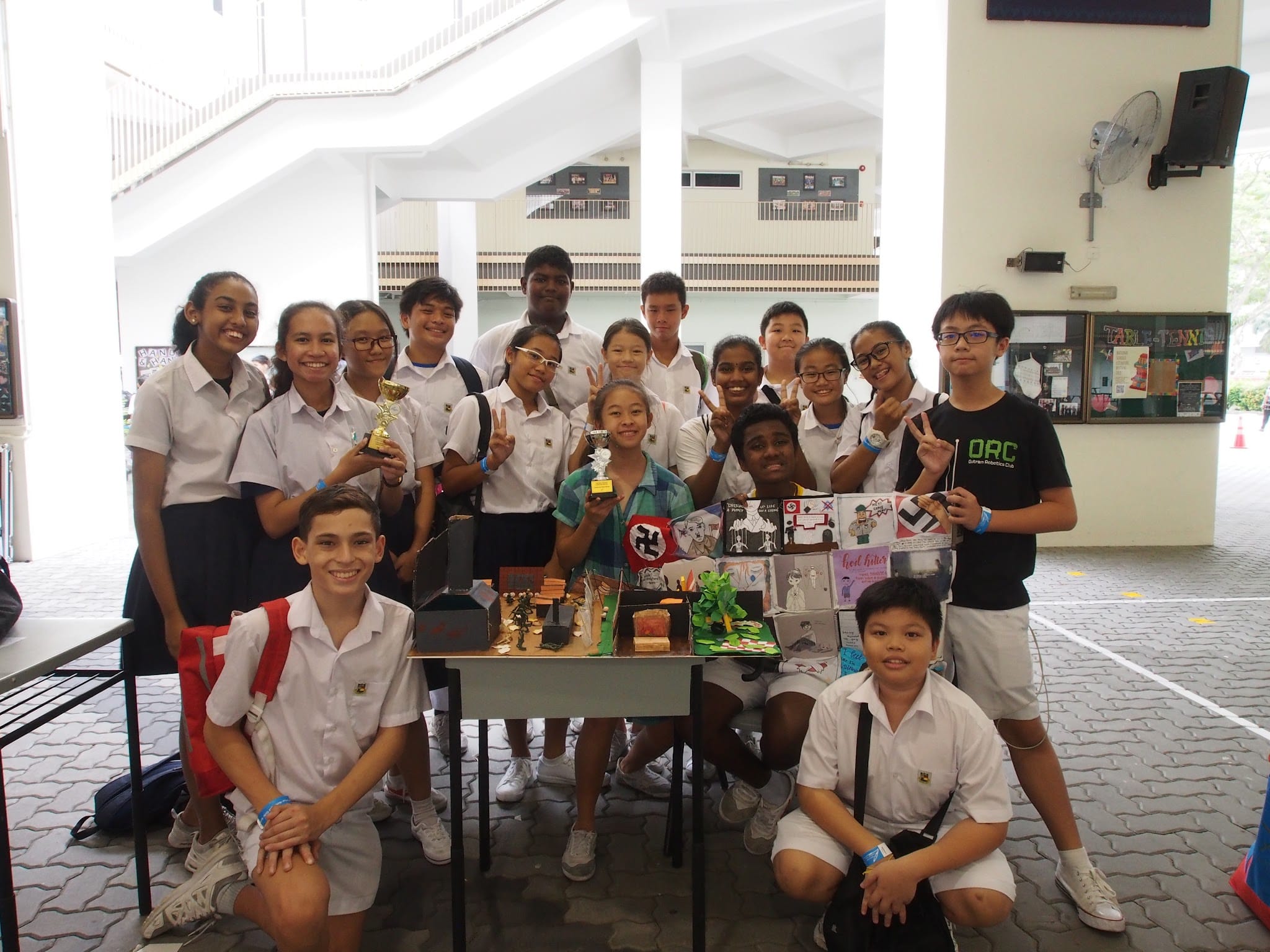 A group of school students in uniforms pose together around a table displaying project models and posters indoors.