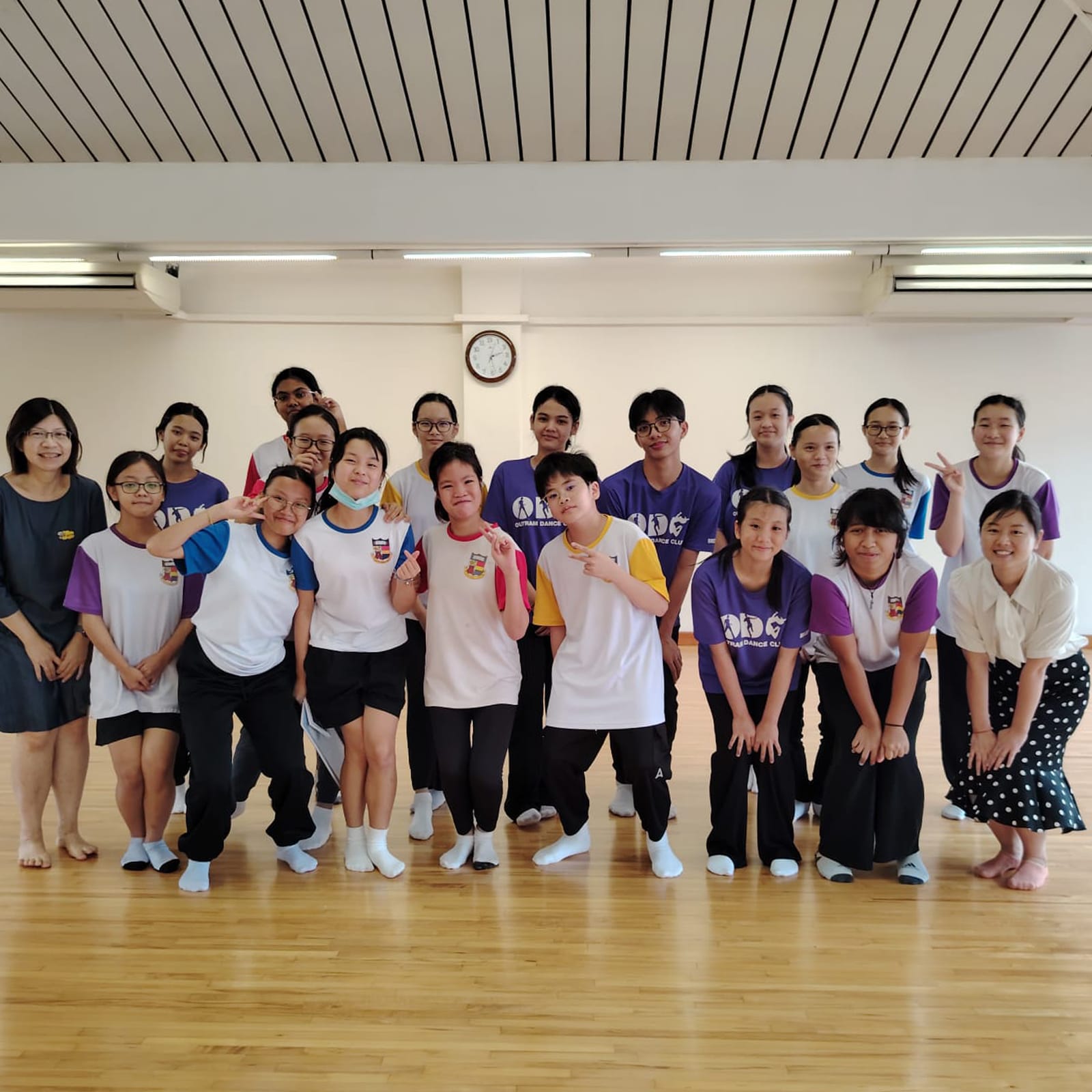 Large group photo of Outram Dance Club members and teachers posing in a dance studio, some kneeling in front, with a wall clock visible behind them.