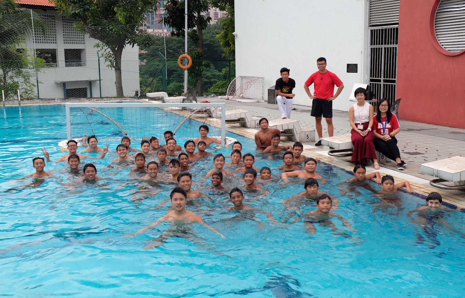 A large group of young boys posing in an outdoor pool with a water polo goal, while four adults stand nearby.