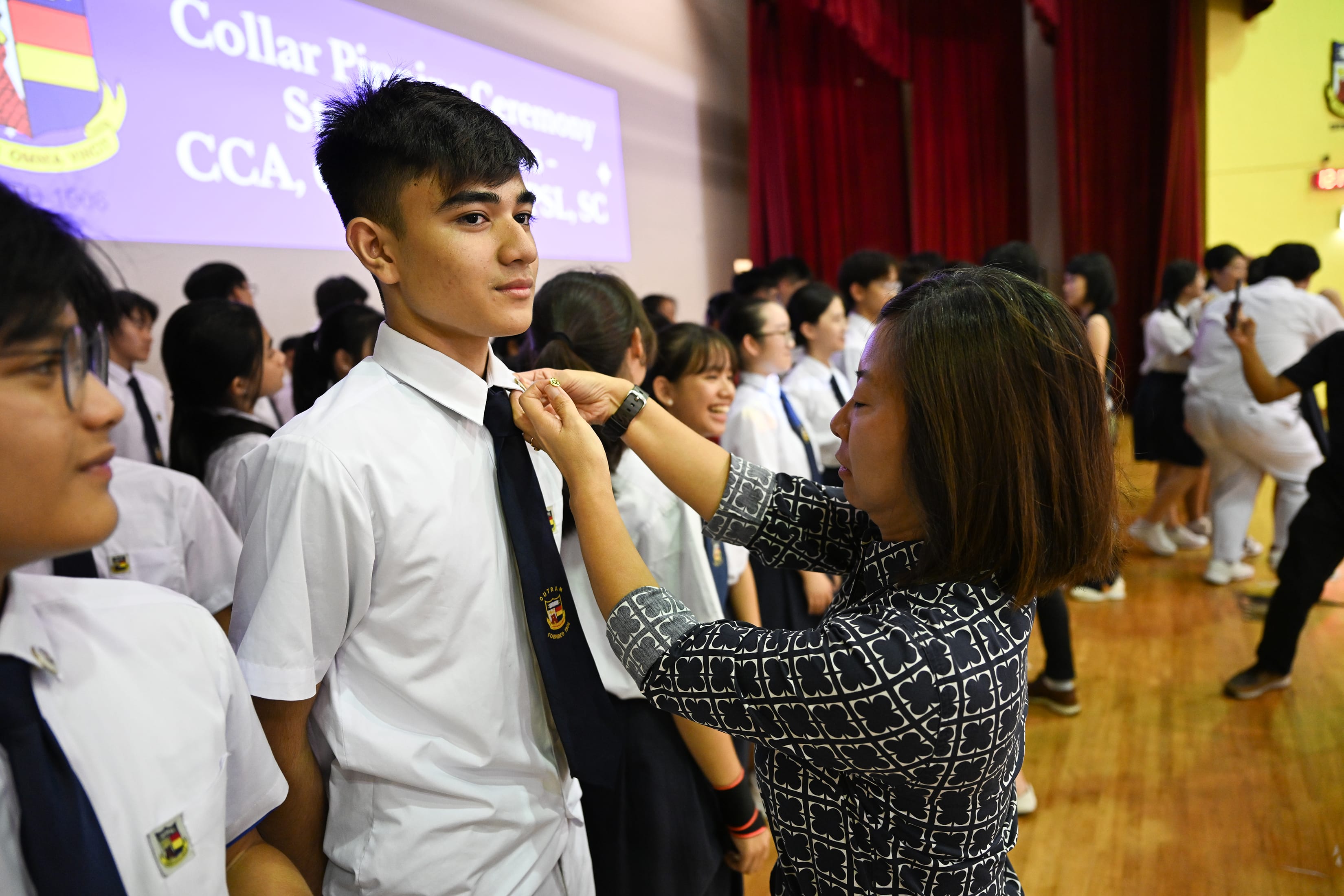 A lady in black and white dress helping a boy in white uniform wear a badge on his collar.