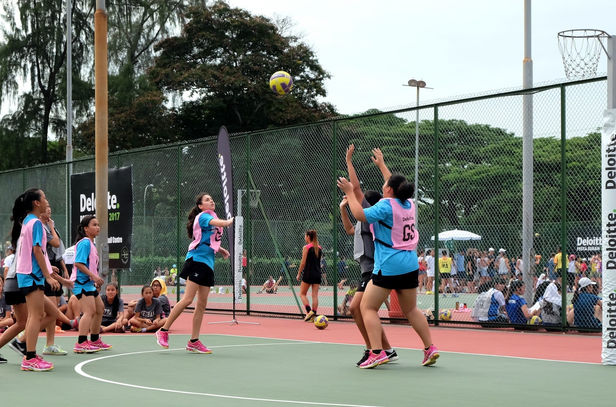Girls in blue and pink bibs play netball on an outdoor court, with a ball in the air near the hoop.