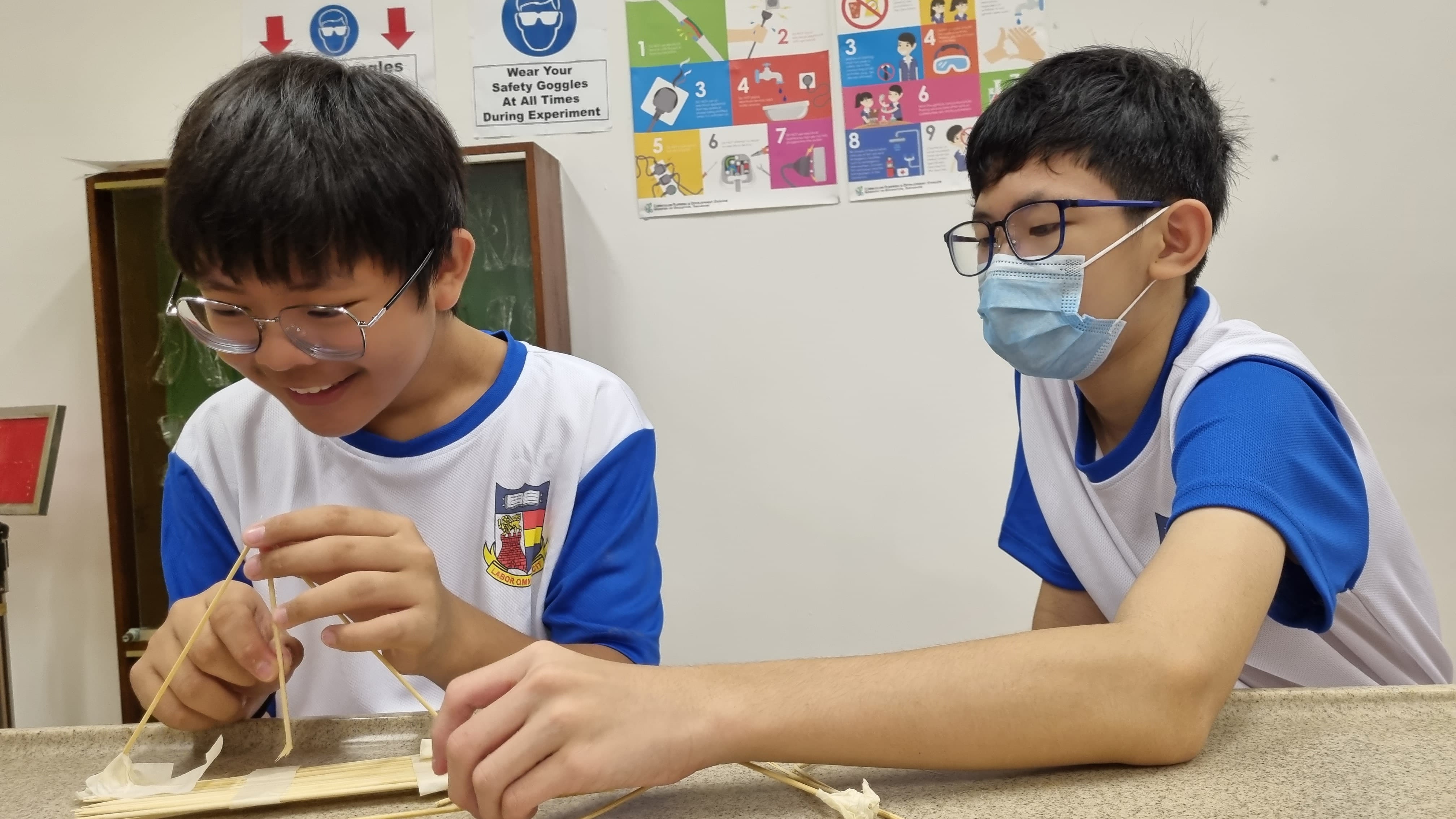 Two boys in school uniforms build a structure with wooden sticks at a desk in a classroom with safety posters.