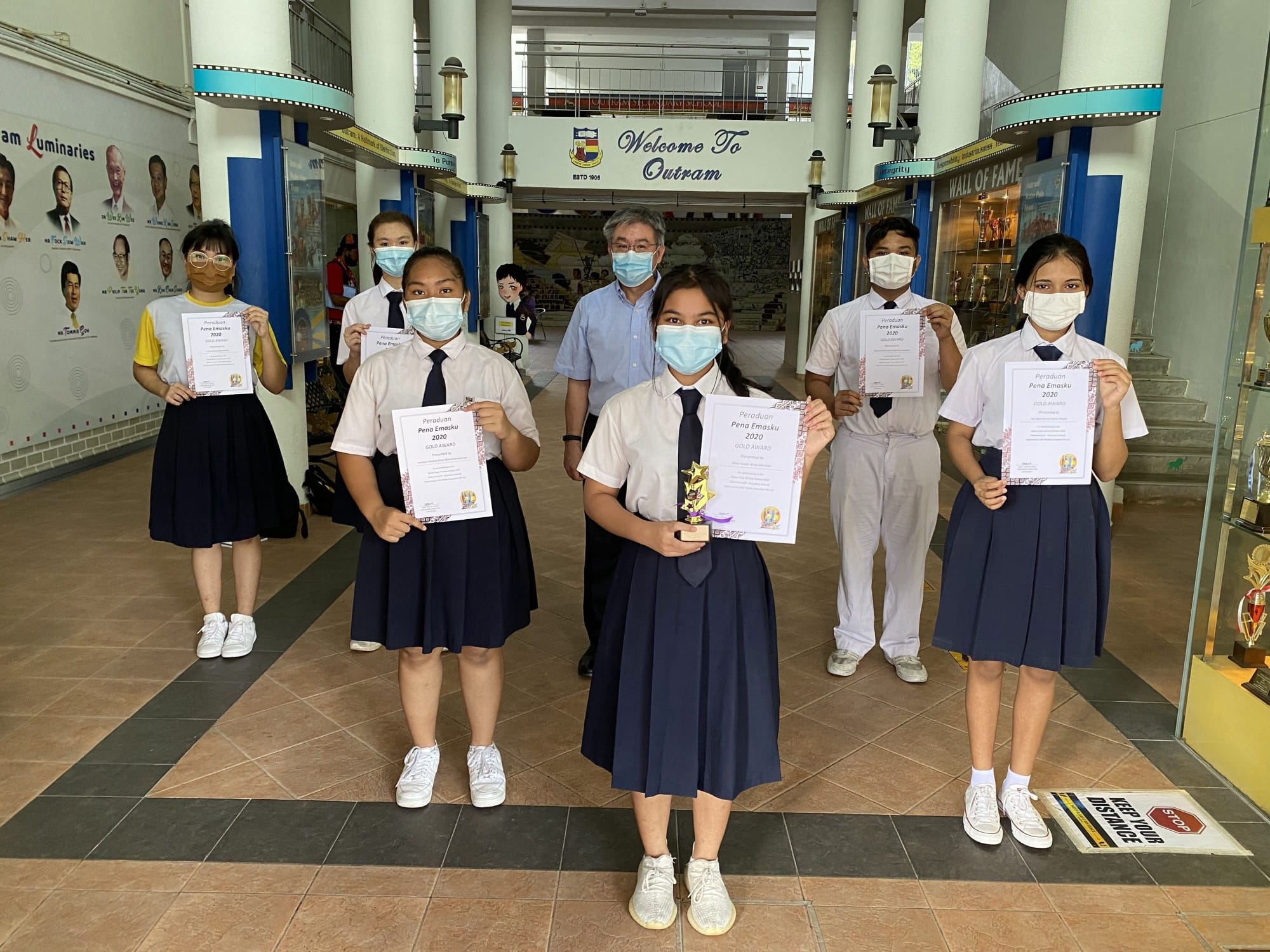 Students in uniforms and masks hold Pena Emasku 2020 award certificates inside Outram Secondary School.