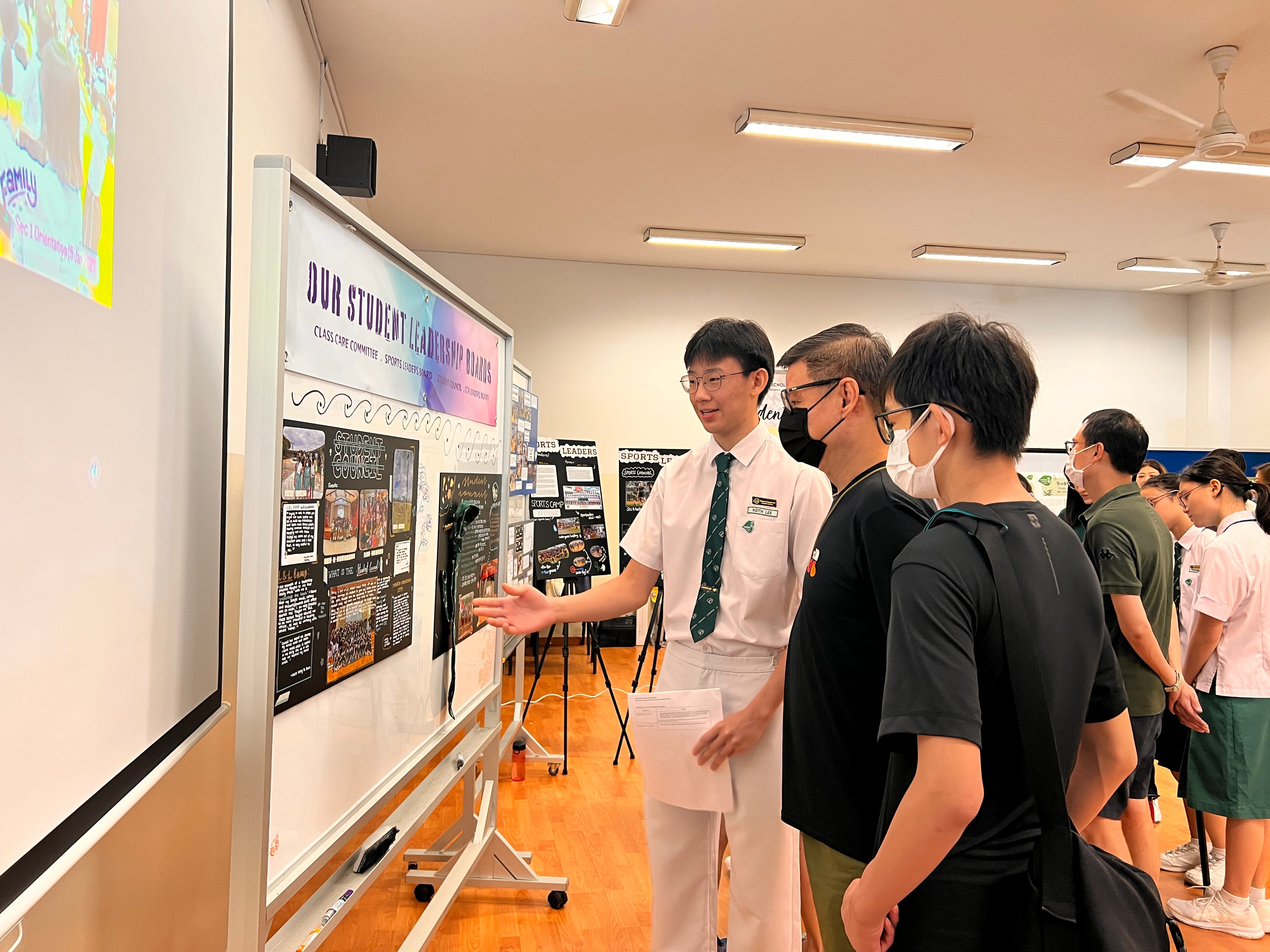 Student in uniform presents a poster board titled Our Student Leadership Boards to a small group indoors.