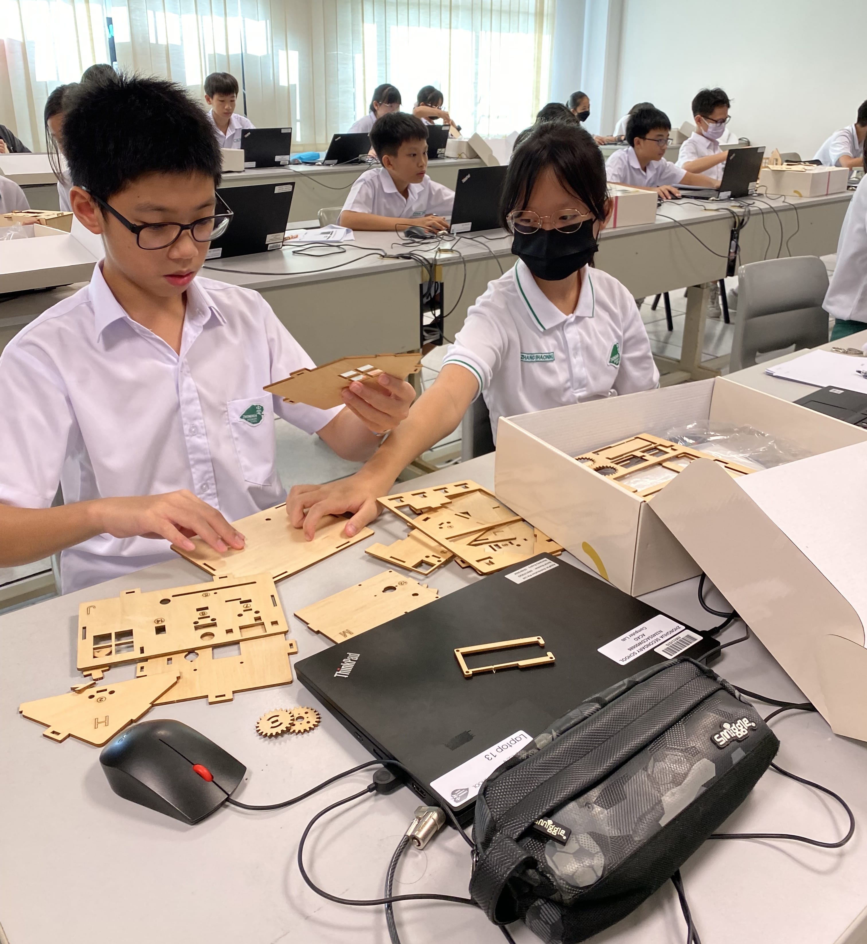 Two students in school uniforms assemble laser-cut wooden puzzle pieces on a desk in a classroom.