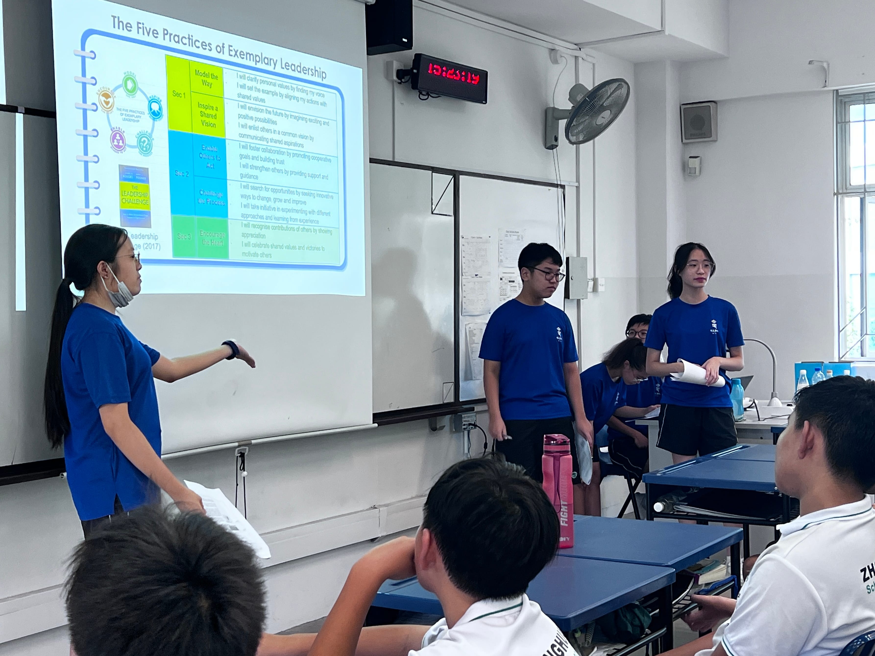 Three students in blue shirts present a slide on Five Practices of Exemplary Leadership to classmates in a classroom.