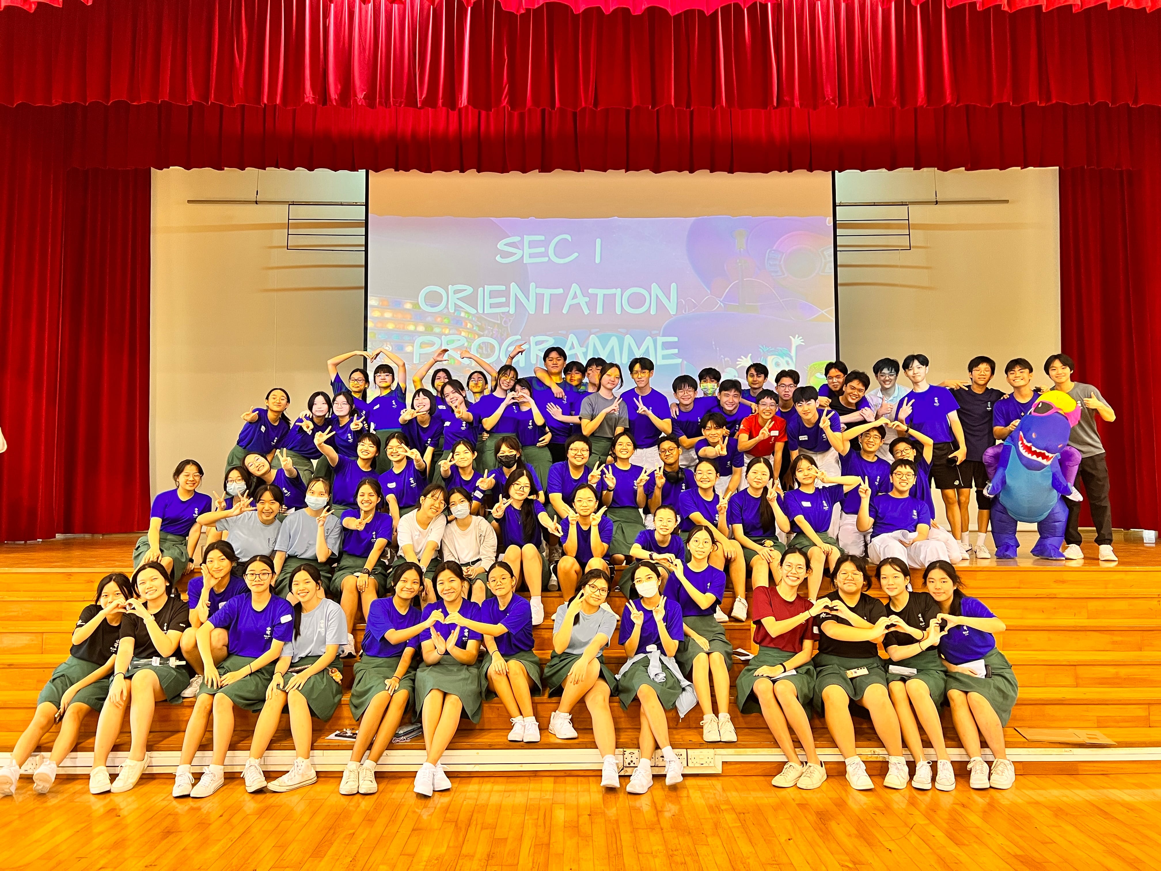 Large group of students posing on a stage under red curtains, with a Sec 1 orientation screen behind.