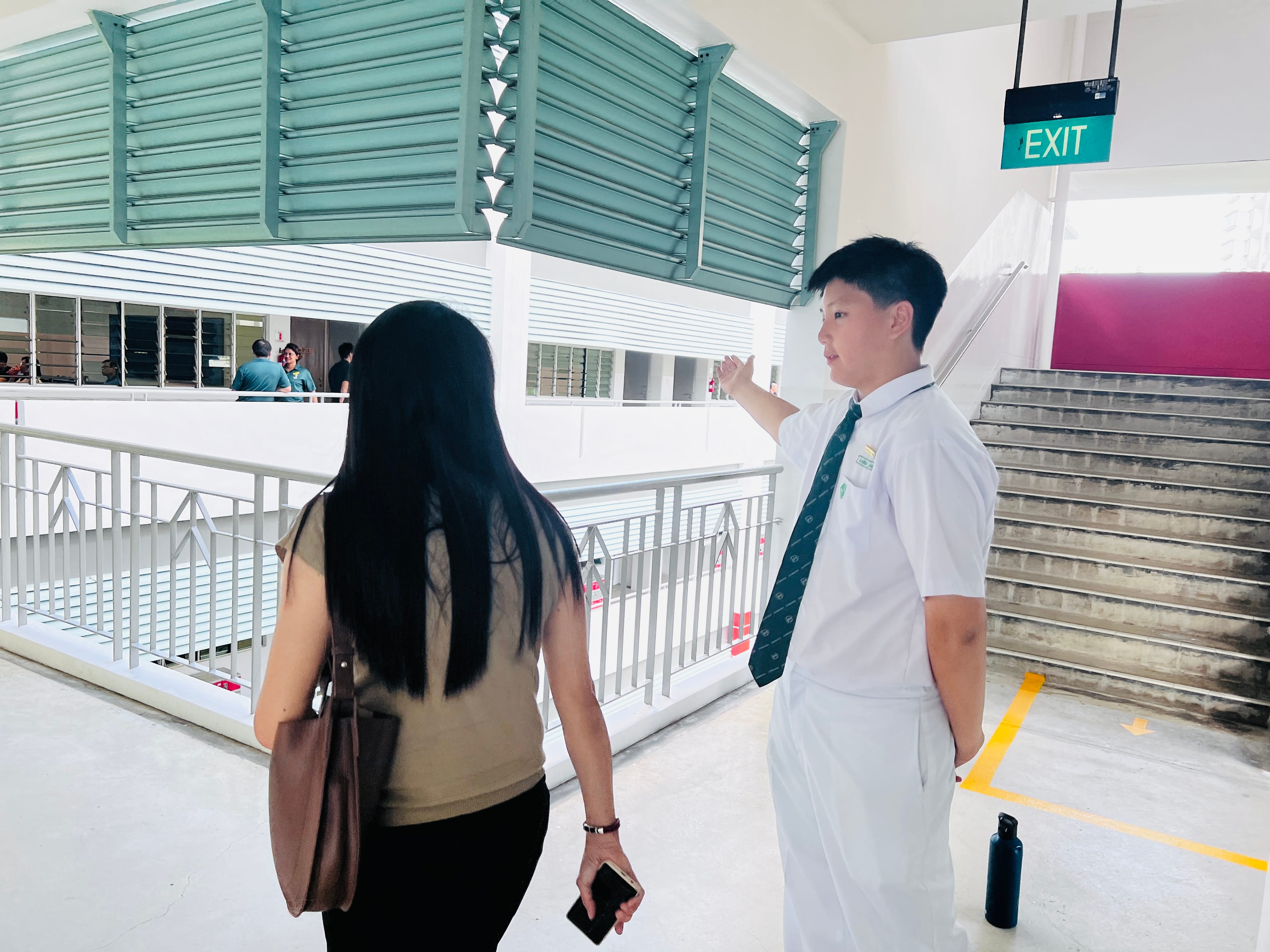 A student in white uniform and tie gestures directions to a woman with long black hair in a school building.