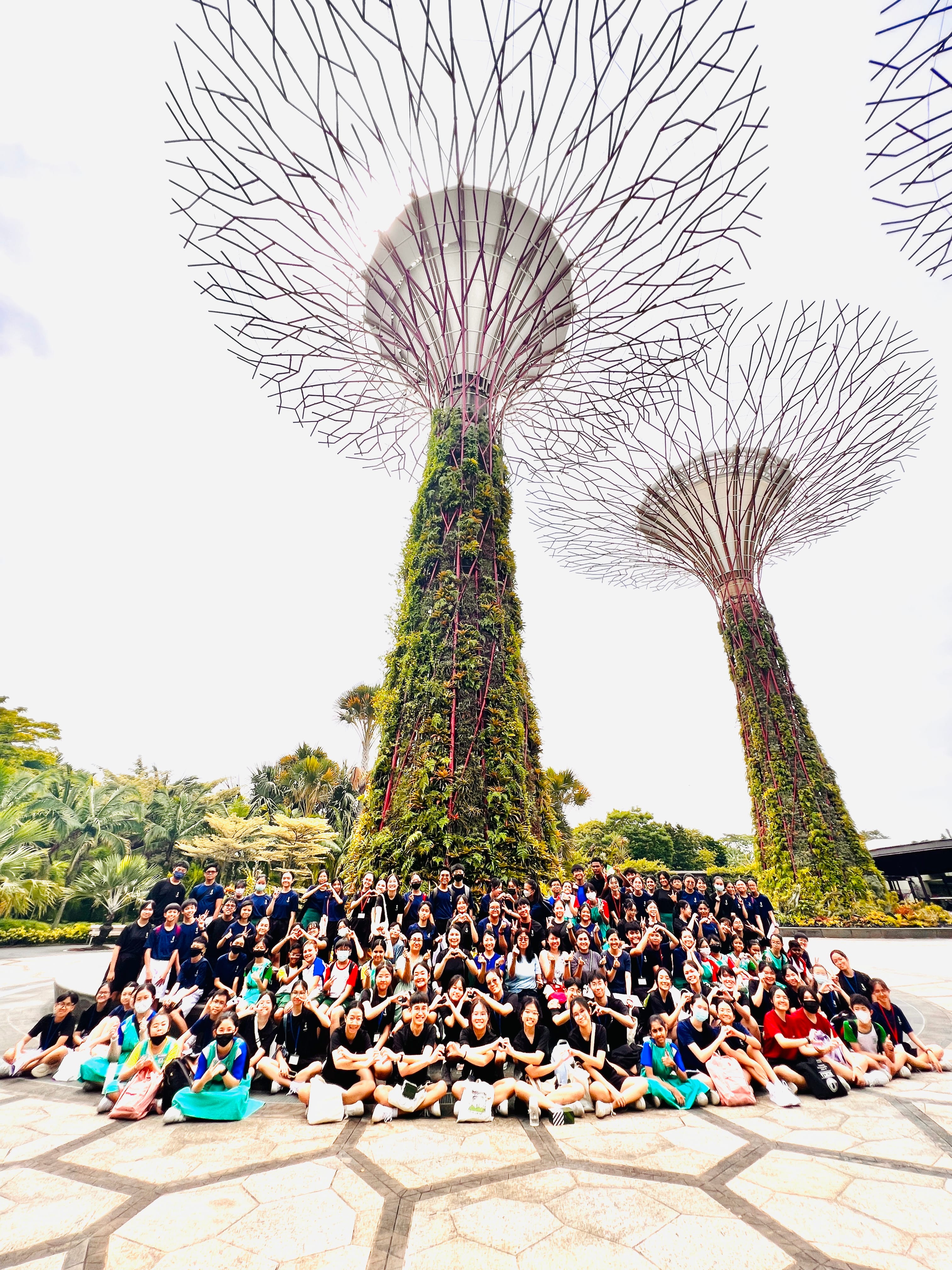 Large group posing in front of two towering tree-like metal structures covered in greenery, viewed upward