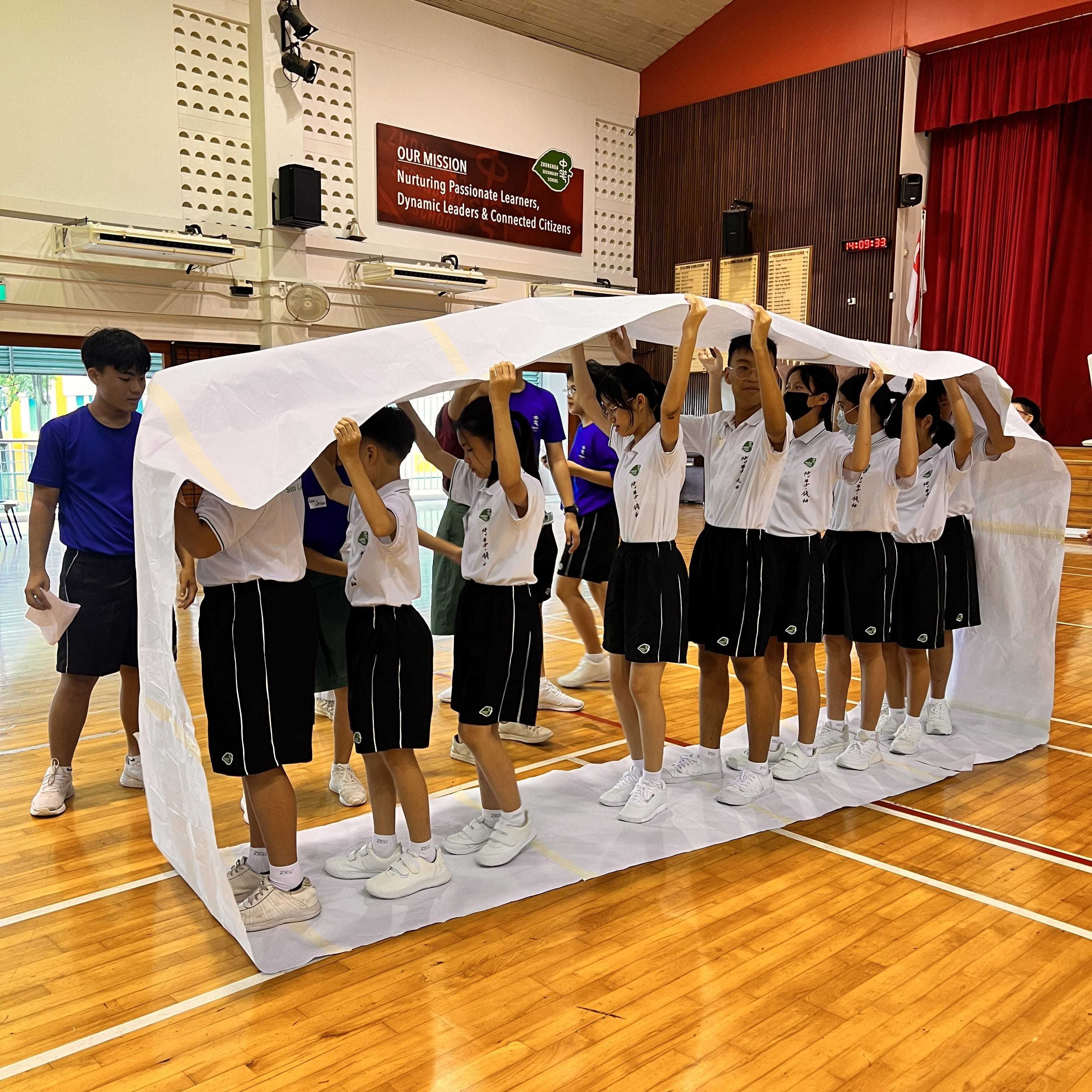 Students in school uniforms hold up a large white sheet over their heads in a school gymnasium.