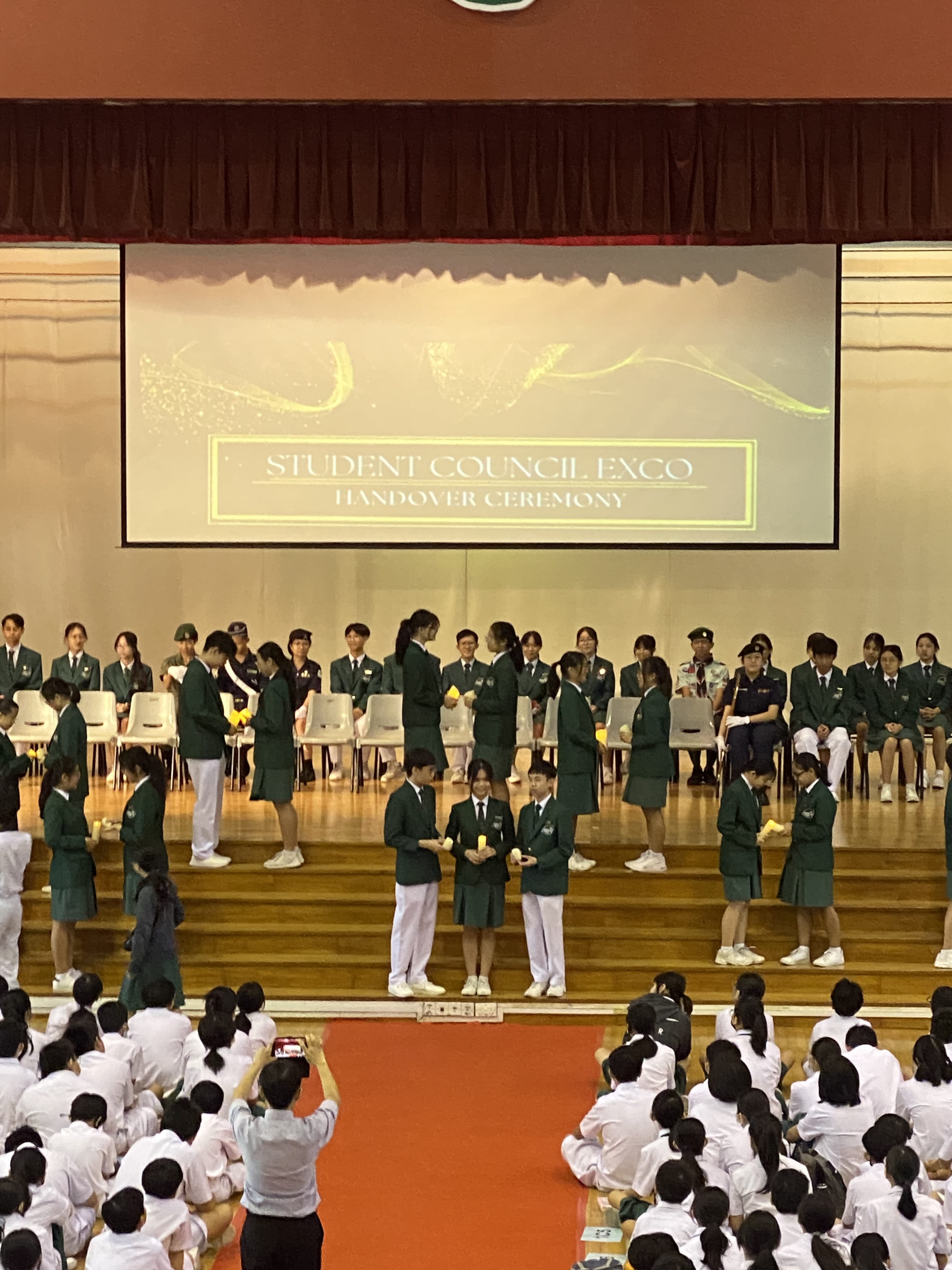 Students in uniforms on a stage during a Student Council Exco Handover Ceremony, with seated audience watching.