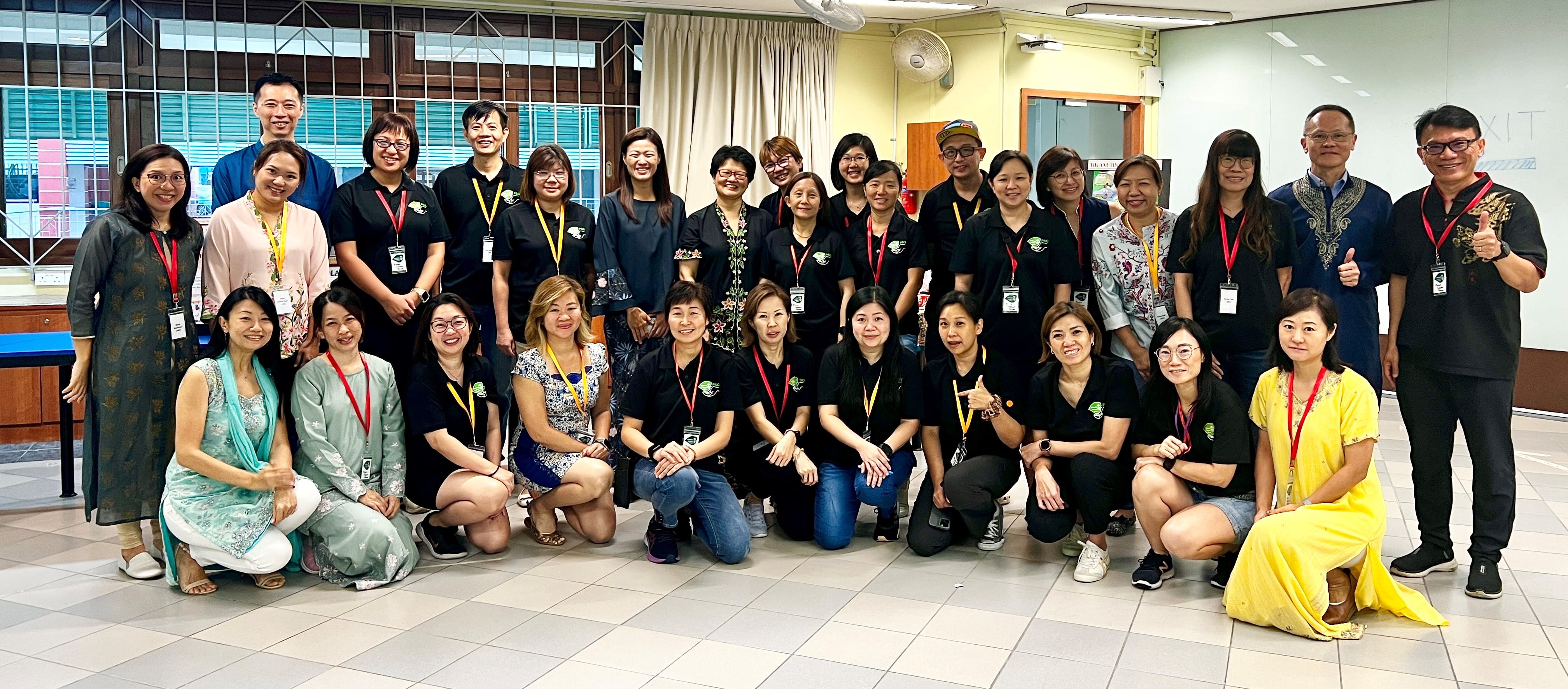 A large group of about 30 adults wearing lanyards pose together indoors for a group photo.