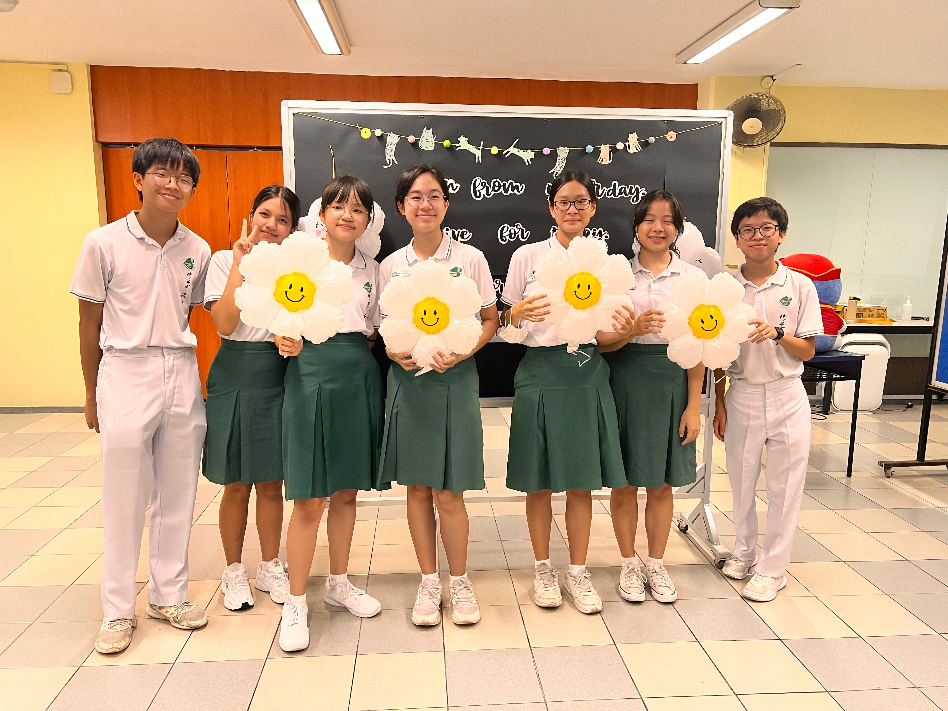 Seven students in white and green uniforms pose in a classroom, holding large smiley-face daisy balloons.