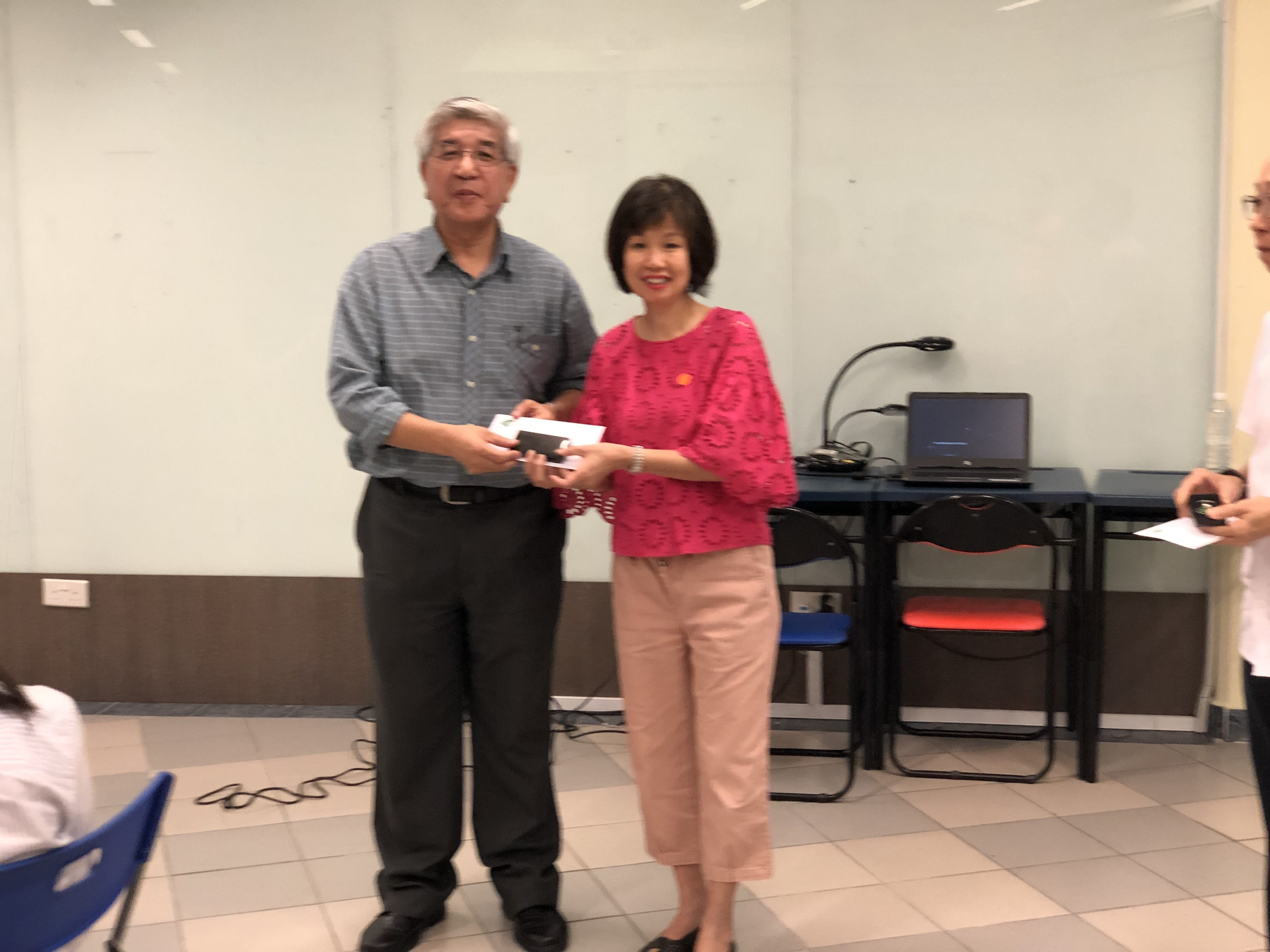 An older man in a gray shirt and a woman in a pink top jointly hold a small box in a meeting room.