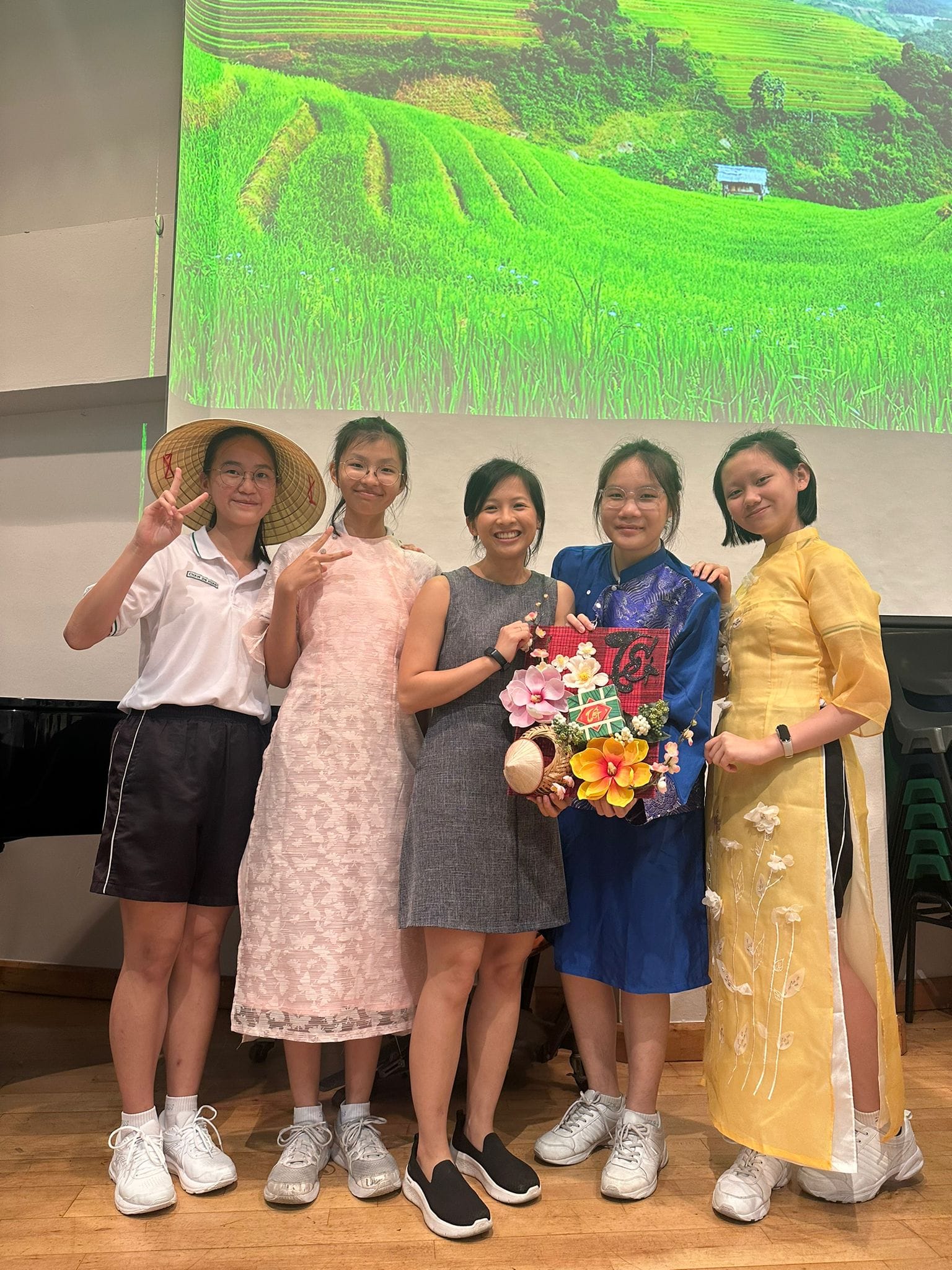 Five young women pose smiling before a projected rice terrace backdrop, some wearing traditional Vietnamese ao dai dresses.