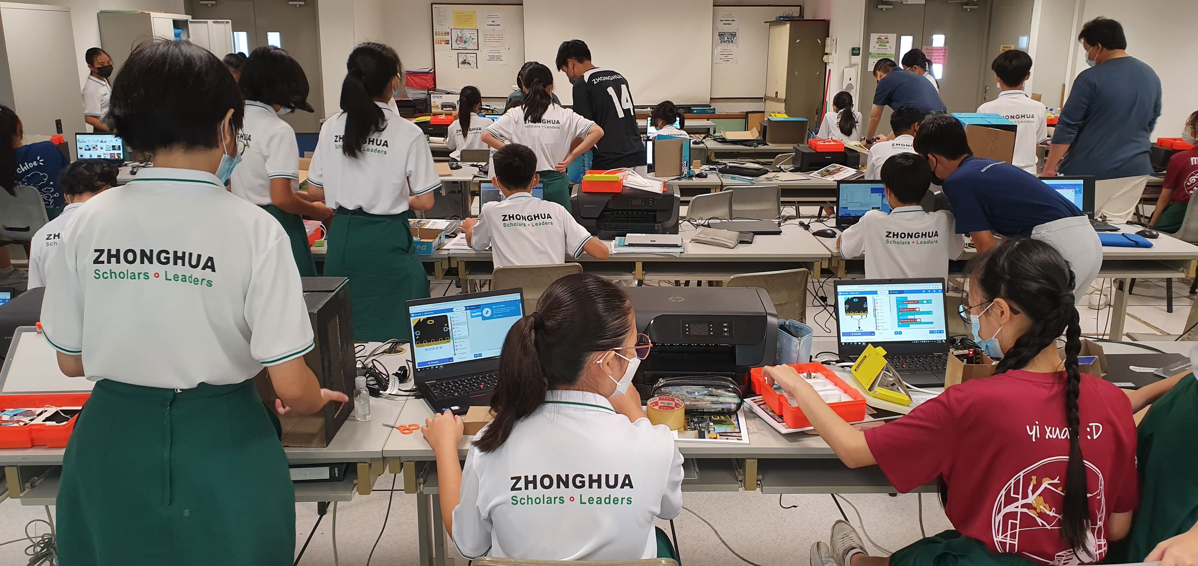 Students in Zhonghua school uniforms work on laptops and printers in a busy classroom workshop.