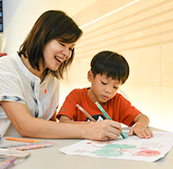 Woman helps boy coloring on paper with markers at a table.