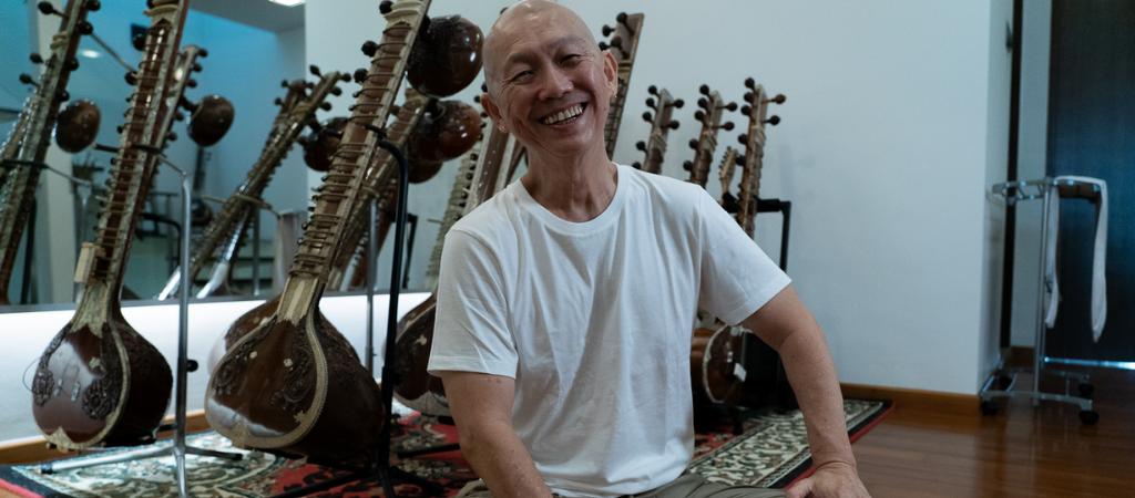 Man in a white t-shirt and glasses, sitting with a sitar, smiling in a music room.