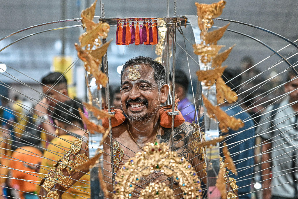 Devotee smiling while carrying a kavadi structure, with hooks and decorations attached to his body.
