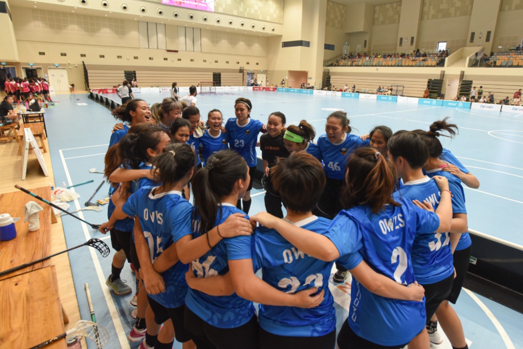Team huddle of female floorballers in blue jerseys and the goalkeeper in black on an indoor floorball court.
