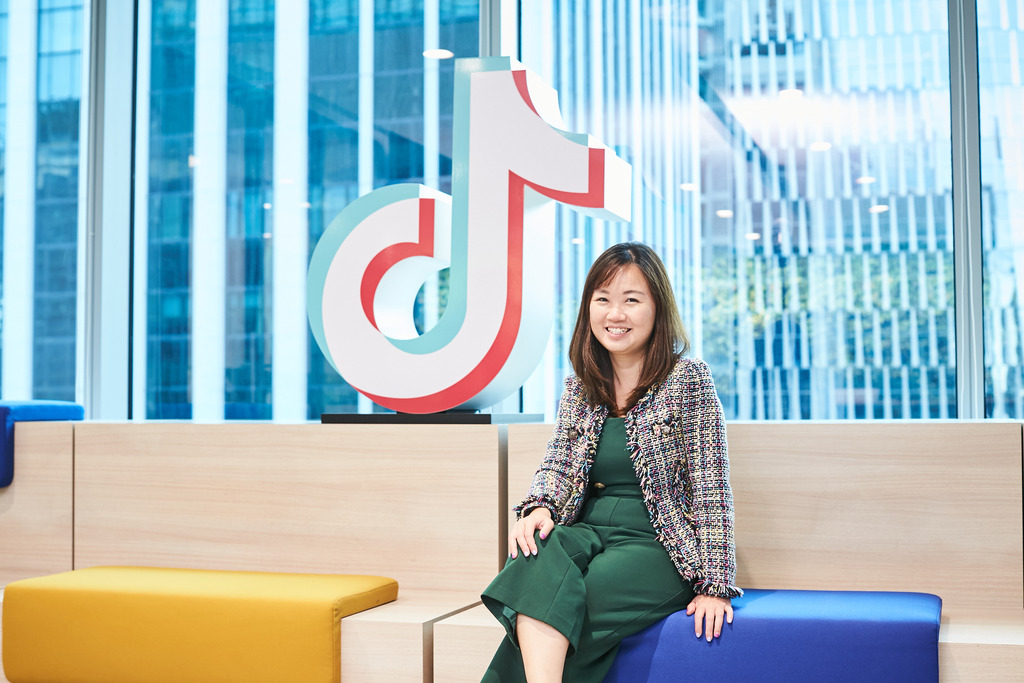 Woman in a green dress sitting on a sofa next to a large TikTok logo in an office setting