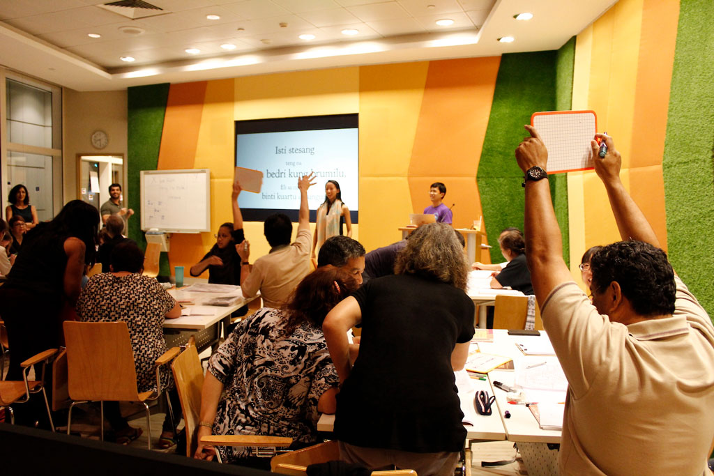 A busy classroom with participants raising hands and a screen showing a learning activity.
