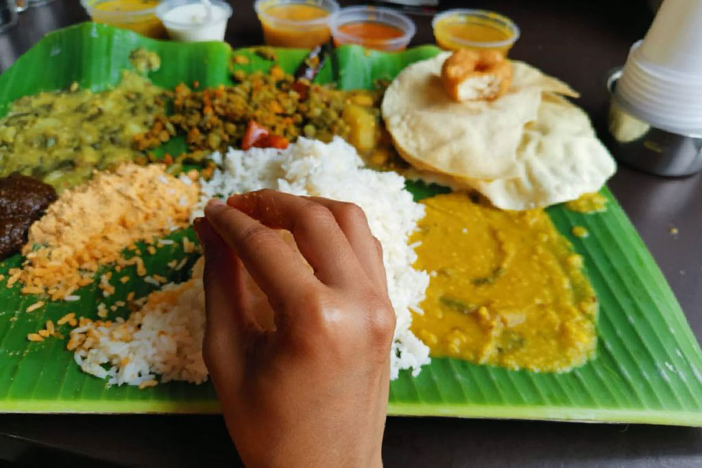Hand reaching for rice on a banana leaf meal with various Indian vegetarian dishes.