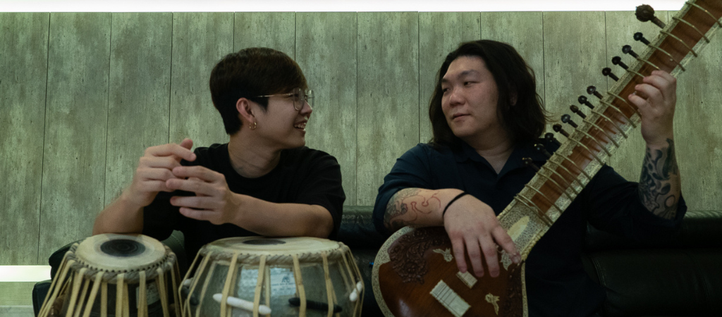 Two musicians sitting side by side, one playing tabla and the other playing sitar, smiling at each other