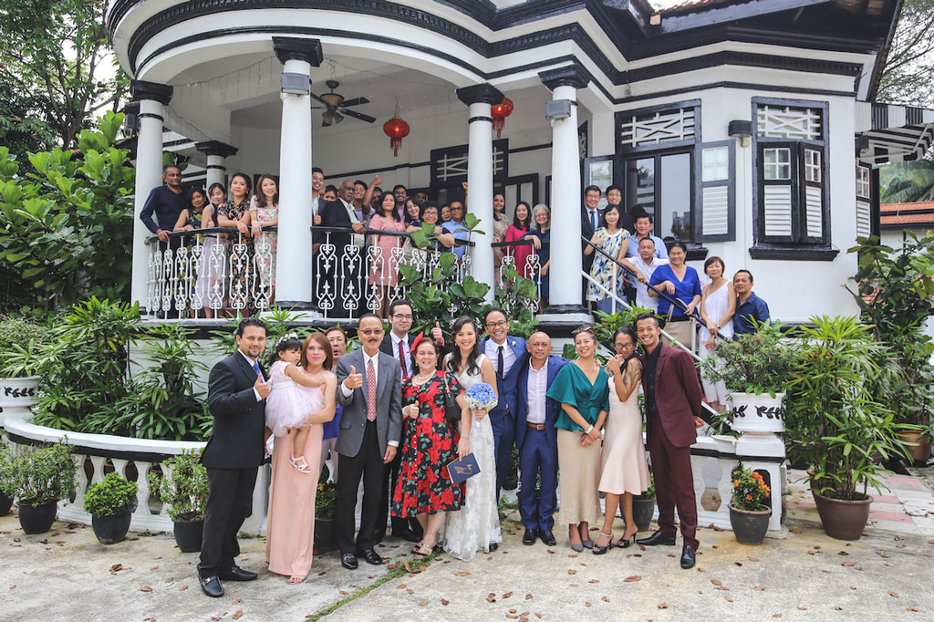A large group of people posing for a photo in front of a traditional house with greenery.