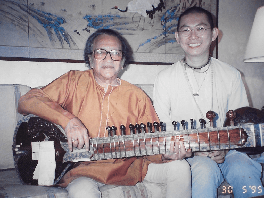 Two men sitting with a sitar, one elderly with glasses, the other wearing a light shirt, smiling together
