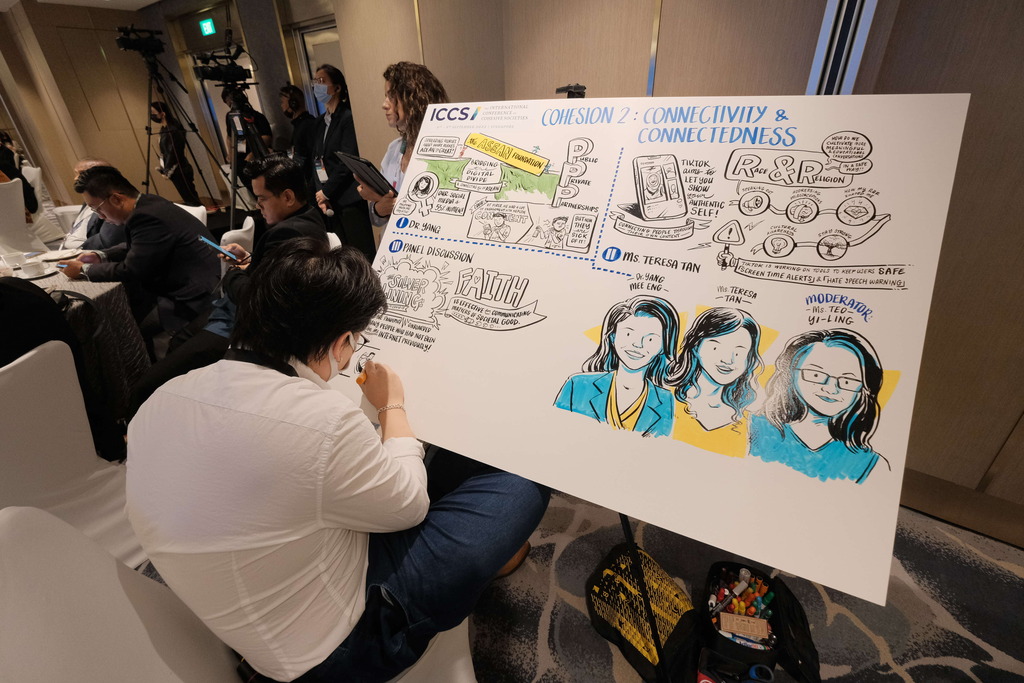 Person drawing illustrations on a large board during a panel discussion, with audience in the background