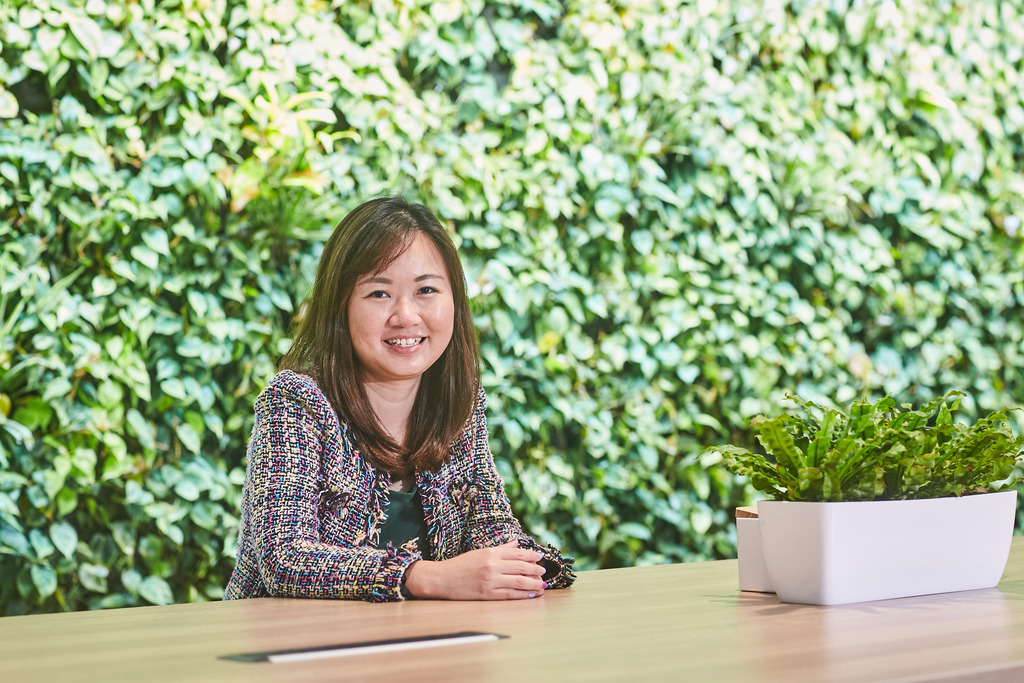 Woman smiling in front of a lush plant wall, seated at a wooden desk