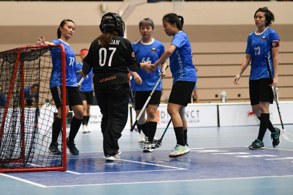 Female floorballers in blue jerseys tapping hands with the goalkeeper in black near the goalpost in the court.