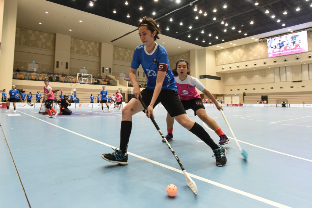 Female floorballer in blue sweeping the ball and shielding it from an opponent in white and pink in a game.